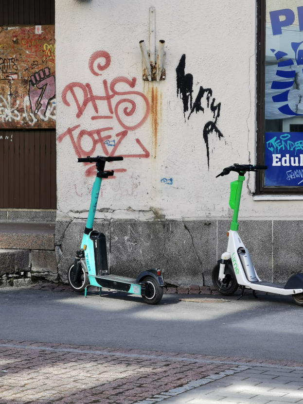 Zwei Elektro-Scooter nebeneinander auf einer Straße geparkt, mit einer Graffiti-bedeckten Wand und einem Plakat an einem Fenster im Hintergrund.