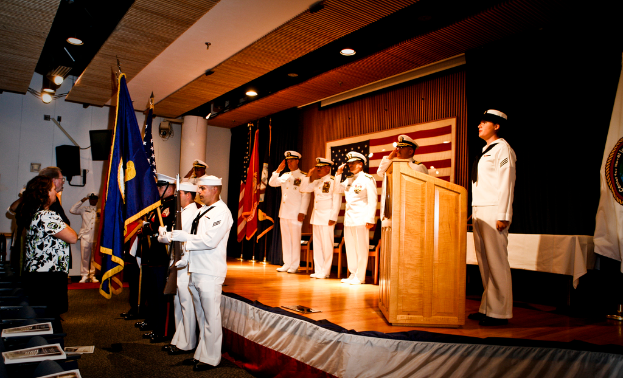 Eine offizielle Veranstaltung mit Menschen, die auf einer Bühne stehen und salutieren, Flaggen in rot, blau und weiß, Stühle, ein Podium und ein Tisch mit einer weißen Decke, sowie ein Fernseher und ein Lautsprecherbox im Hintergrund.