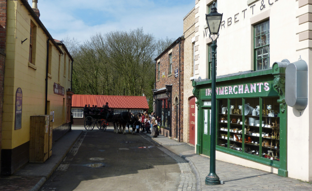 Eine belebte Straßenszene mit einem Pferdewagen, der Menschen transportiert, Fußgängern auf den Gehwegen, Gebäuden mit Fenstern und Schildern, einer Straßenlaterne und Bäumen und Himmel im Hintergrund.