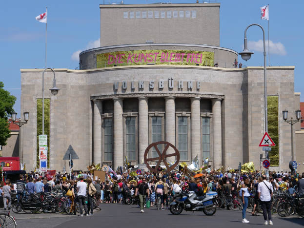 Große Gruppe von Menschen versammelt sich vor einem Gebäude mit Säulen und Text in Berlin, Deutschland, mit Motorrädern, Fahrzeugen, Straßenschildern und Bäumen im Hintergrund.