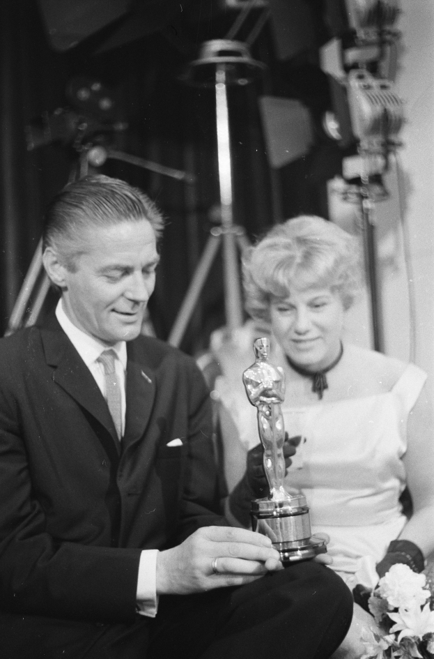 Ein Mann in einem schwarzen Blazer, einem weißen Hemd und einer Krawatte h├Ąlt eine Oscar-Statue, waehrend er neben einer Frau in Abendgarderobe sitzt, die einen Blumenstrau├č h├Ąlt, mit Hintergrundbeleuchtung in einem Schwarz-Wei├č-Bild.