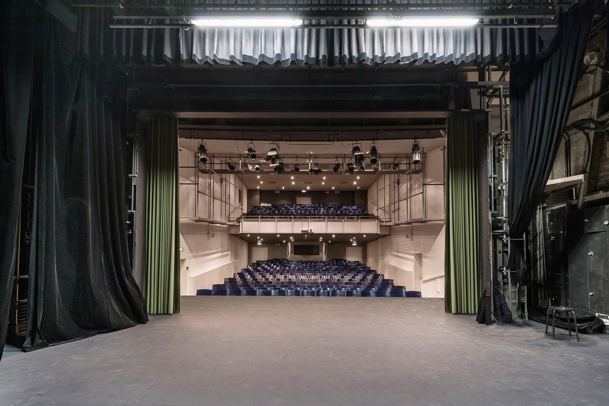 Großer Zuschauerraum mit schwarzen Vorhängen, einer Bühne, Deckenlampen und Stühlen, mit Gegenständen auf der rechten Seite.