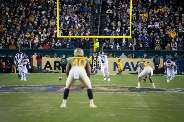 Ein Fussballspiel im Gange mit Spielern in Sportbekleidung und Helmen im Vordergrund und einer Zuschauermenge im Hintergrund, mit sichtbaren Pfosten und Texttafeln.