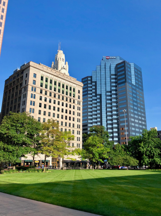 Ein Stadtpark mit hohen Gebäuden im Hintergrund, umgeben von grünem Gras, Bäumen und Laternen, mit Menschen, die herumlaufen und dem Himmel über ihnen.