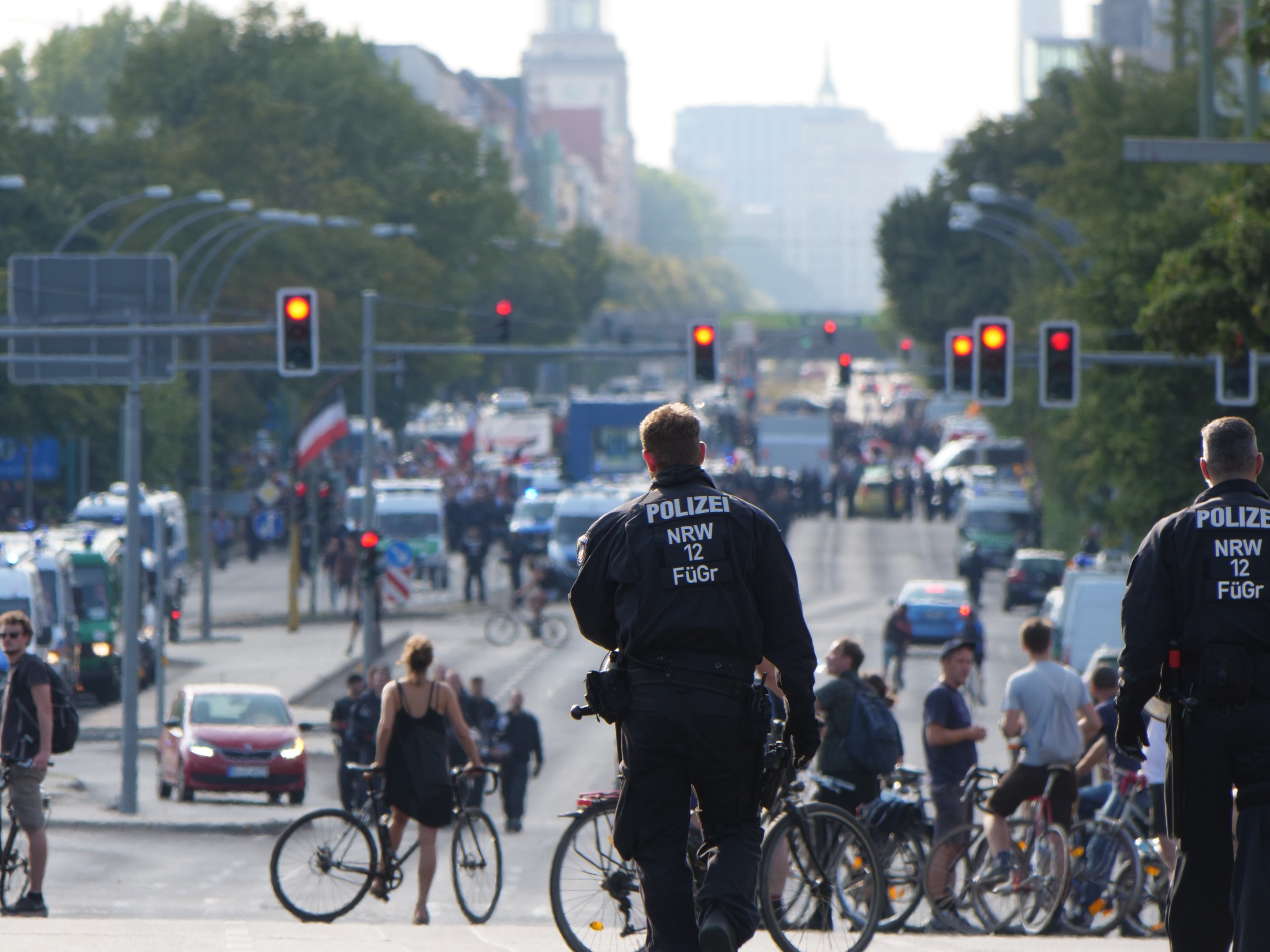 Zwei Polizisten neben Radfahrern auf einer Straße mit Fahrzeugen, Verkehrszeichen, Bäumen, Gebäuden und einem klaren Himmel.