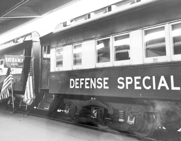Schwarz-weißes Foto eines Zuges mit der Aufschrift "Defense Special" auf den Gleisen mit zwei Flaggen links und einer Decke oben.