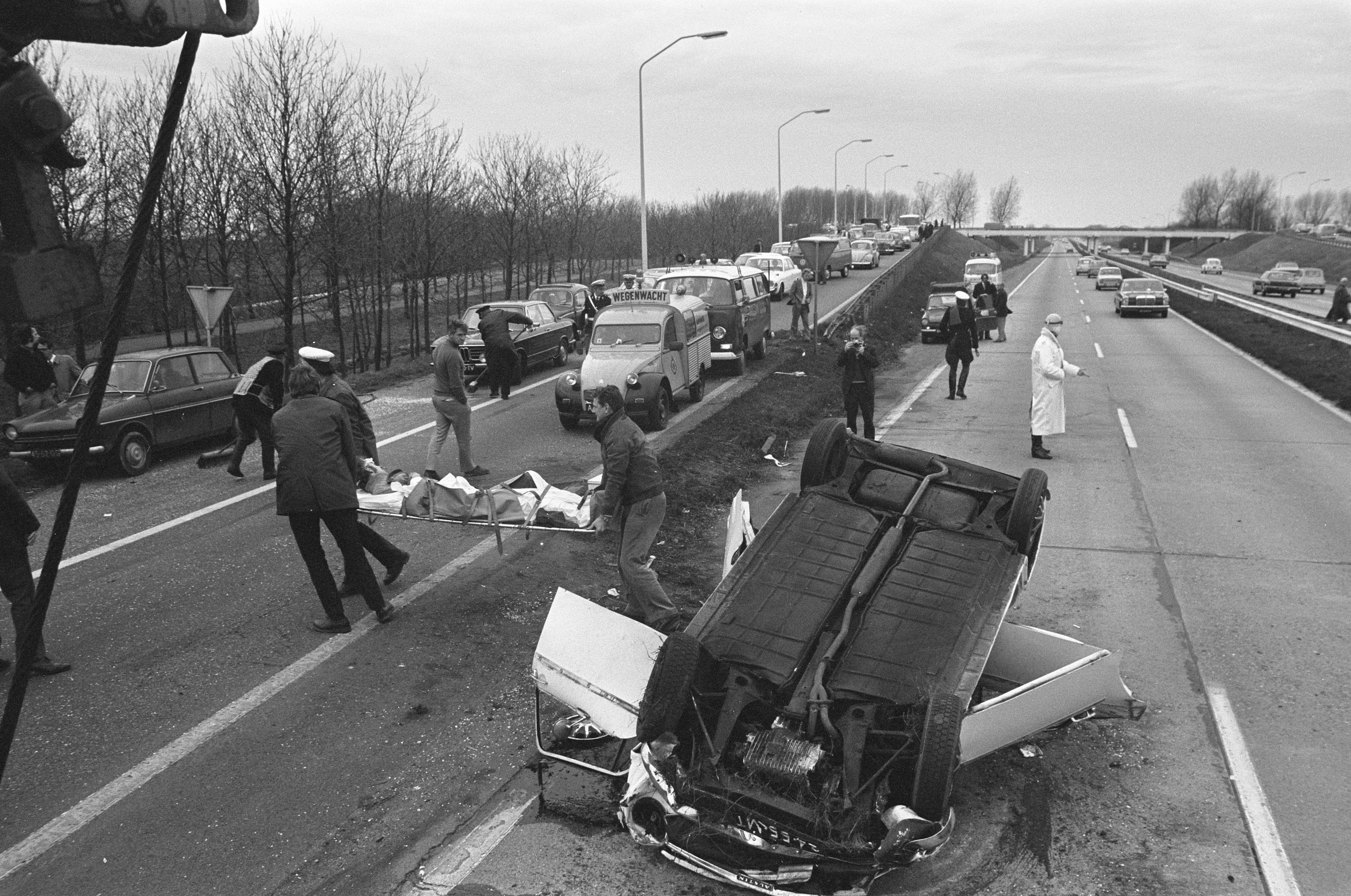 Schwarzes und weißes Bild eines umgestürzten Autos am Straßenrand mit beschädigten Fahrzeugen in der Nähe und einer Gruppe von Menschen, die darum herumstehen, vor dem Hintergrund von Laternen, Bäumen, einer Brücke und dem Himmel.