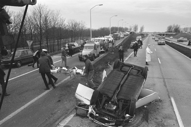 Schwarzes und weißes Bild eines umgestürzten Autos am Straßenrand mit beschädigten Fahrzeugen in der Nähe und einer Gruppe von Menschen, die darum herumstehen, vor dem Hintergrund von Laternen, Bäumen, einer Brücke und dem Himmel.