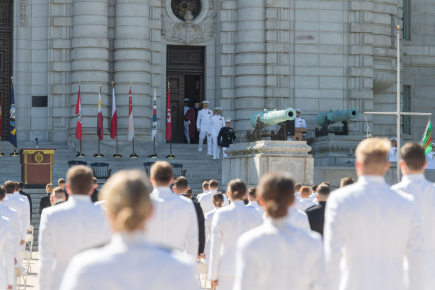 Gruppe von Menschen in weißen Marineuniformen auf einer Treppe vor einem Gebäude mit Säulen, Fahnen, einem Podium und Kanonen während einer Abschlusszeremonie.