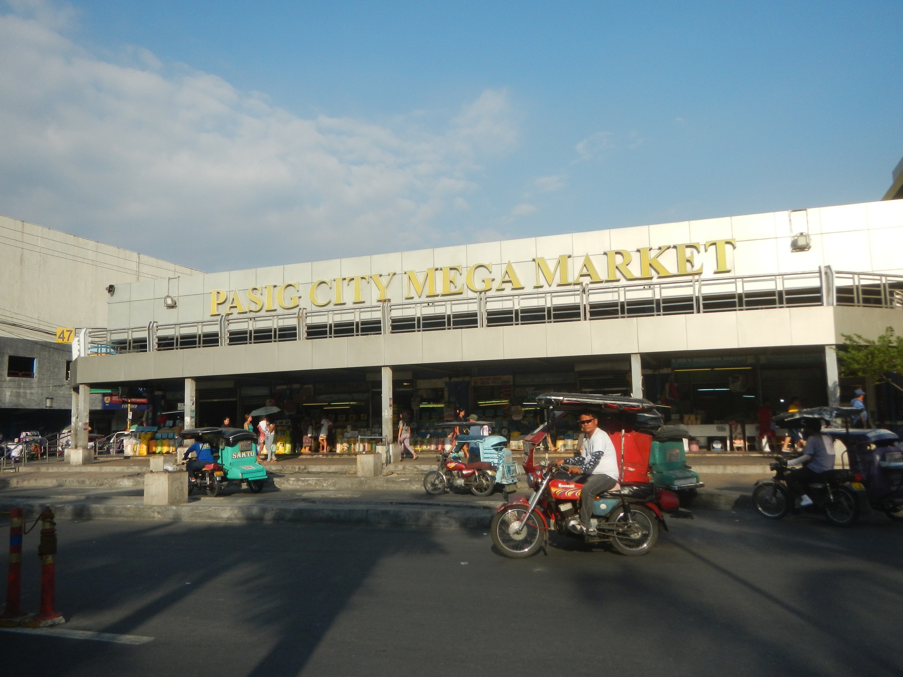 Eine belebte Straßenszene am Pasig City Mega Market mit Fahrzeugen und Fußgängern, ein Gebäude mit sichtbarem Text im Hintergrund und eine bewölkte Himmel.