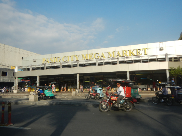 Eine belebte Straßenszene am Pasig City Mega Market mit Fahrzeugen und Fußgängern, ein Gebäude mit sichtbarem Text im Hintergrund und eine bewölkte Himmel.