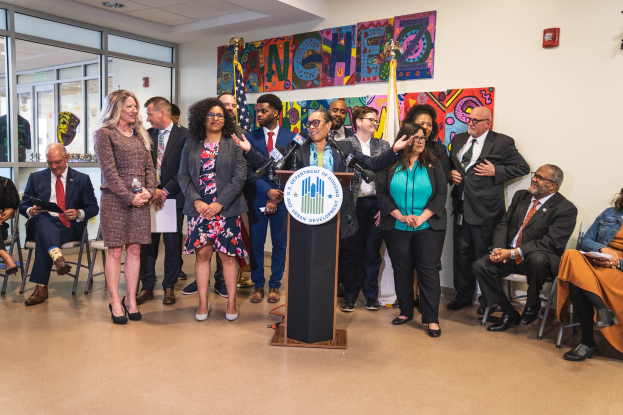 Gruppe von Menschen vor einem Rednerpult mit Mikrofon und einer Tafel mit der Aufschrift "Sancho Stadtrat tritt der Stadtverwaltung bei", mit sitzenden und stehenden Teilnehmern, Flaggen, Feuerlöscher und Deckenleuchten im Hintergrund.