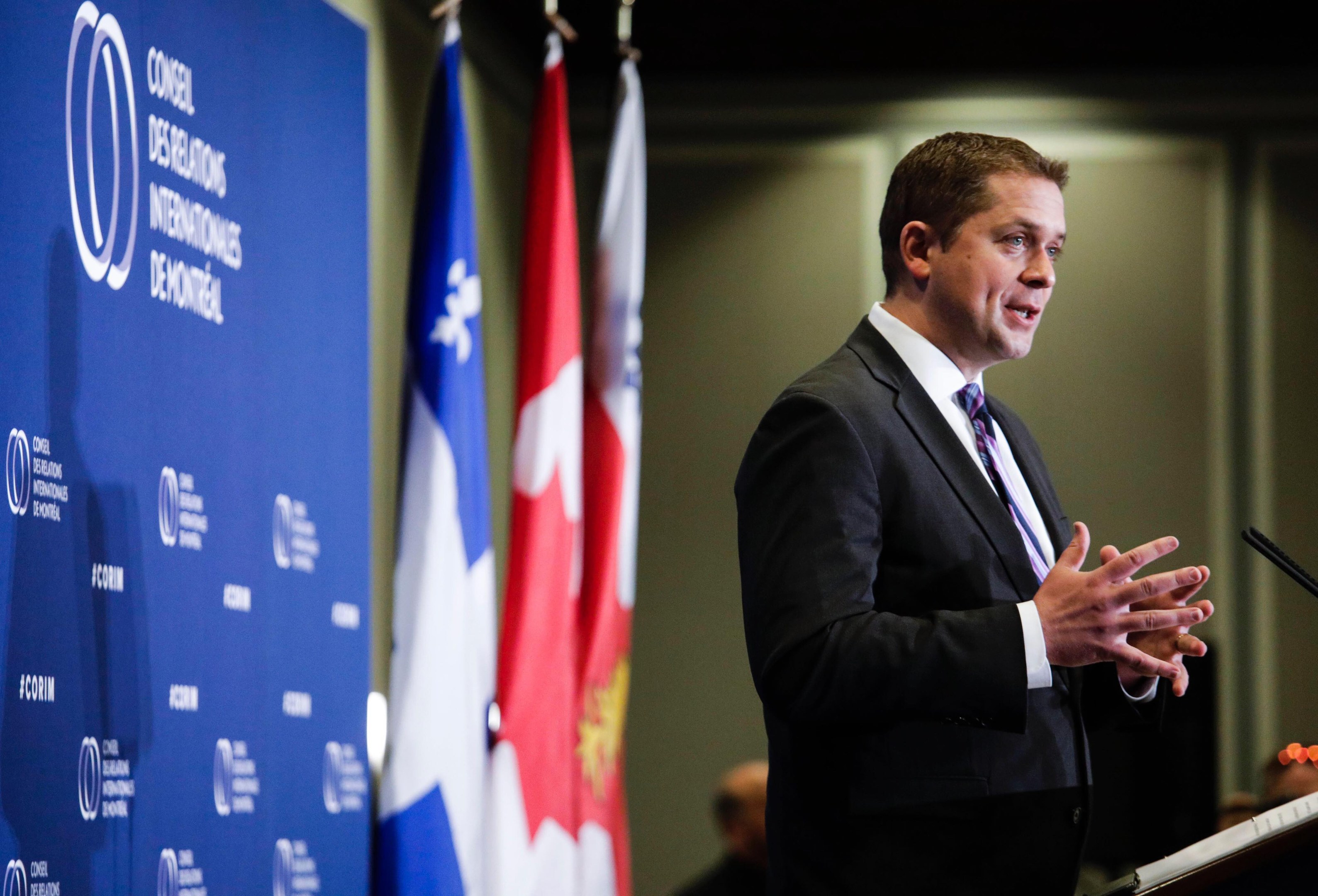 Der kanadische Premierminister steht an einem Rednerpult mit einem Mikrofon, hält eine Rede vor Fahnen und einer Banner, mit Menschen im Hintergrund.