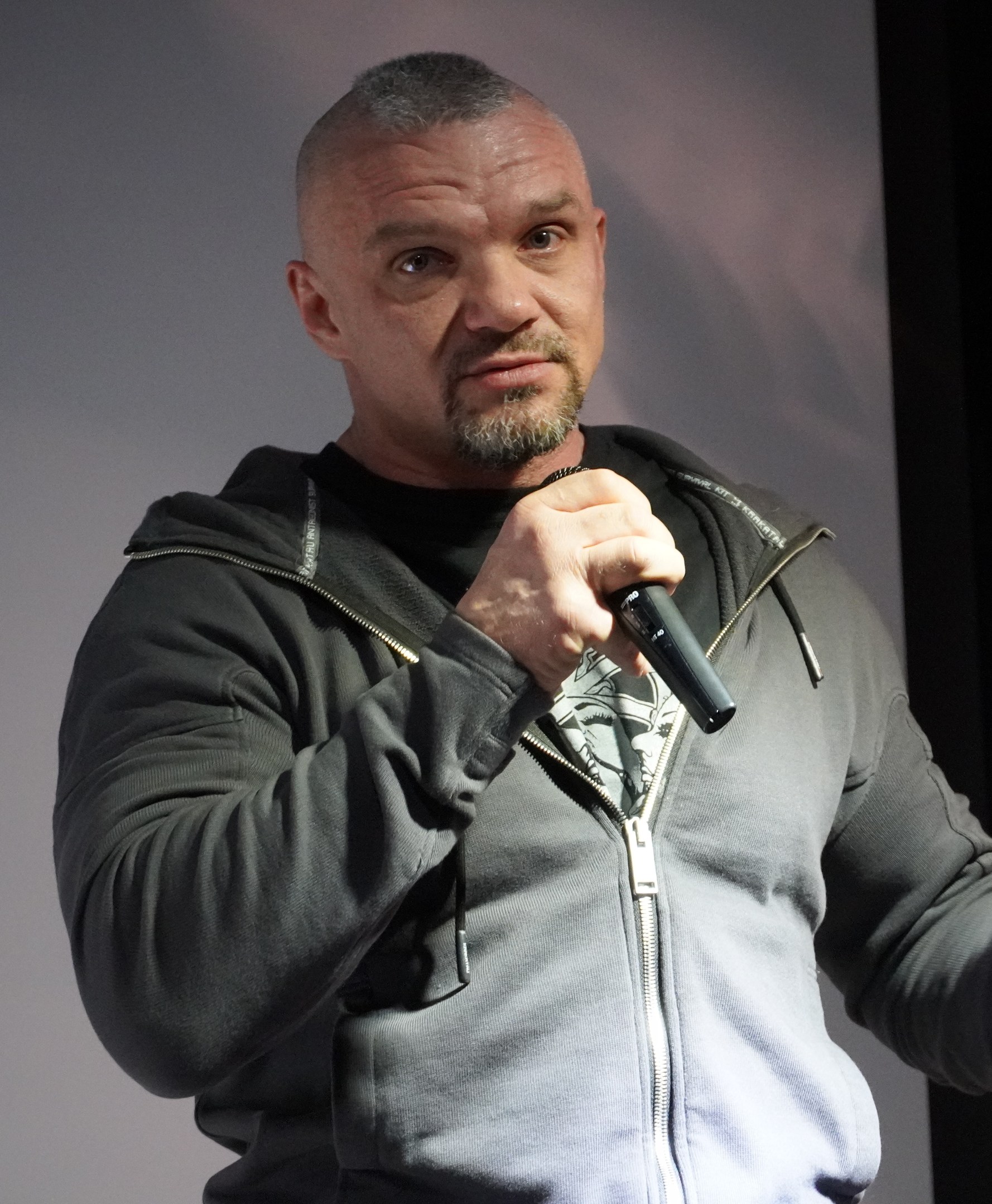 Ein Mann in einer schwarzen Kapuzenjacke und einem schwarzen Anzug mit einem weißen Hemd hält ein Mikrofon und steht selbstbewusst vor einer Wand, wobei er wie ein Komiker aussieht.