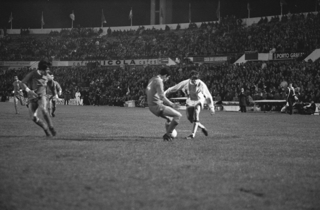 Schwarzes und weißes Bild von Männern, die auf einem Feld in einem Stadion mit Zuschauern in den Rängen und Fahnen im Hintergrund Fußball spielen.
