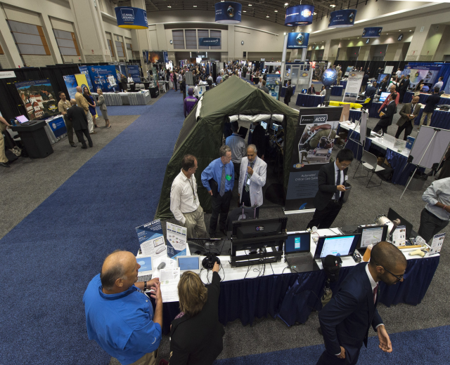 Große Gruppe von Menschen auf einer Karriere-Messe um Tische mit Laptops, Monitoren und Papieren versammelt, mit einem Zelt, Schildern mit Text, Deckenleuchten und Fenstern im Hintergrund.