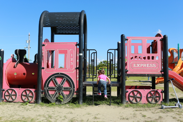 Ein Kind steigt die Stufen von Spielgeräten hoch, mit Bäumen, Polen, Stromleitungen und einer bewölkten Himmel im Hintergrund und einem Roller auf dem Boden rechts.