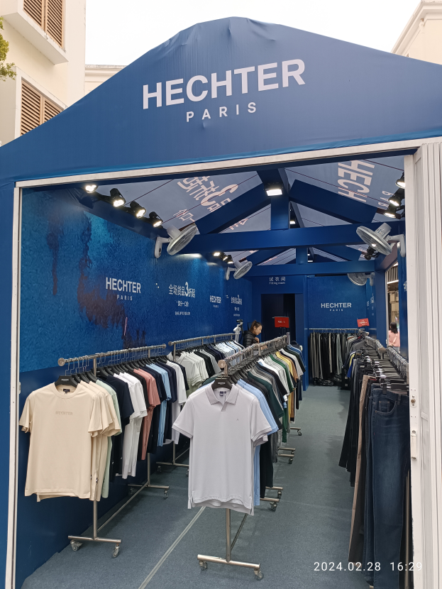 Ein Hechter Paris-Stand auf der Paris Fashion Week, mit Körben an Hängern, einer Person im Stand, Lichtern, einem Gebäude im Hintergrund, einem Baum links und einem sichtbaren Himmel öber dem Bild, mit einem Wasserzeichen in der rechten unteren Ecke.