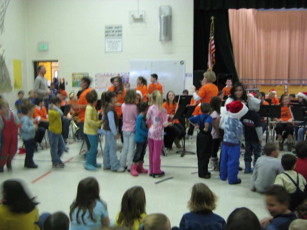 Kinder in bunten Kältern unten auf dem Bild, mit einer Bühne, Vorhang, weißer Tafel, Plakaten, Tür, Schilder, eine Flagge und ein Objekt an der Wand im Hintergrund.