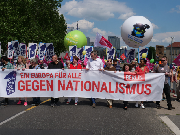 Eine Gruppe von Menschen marschiert auf einer Straße in Berlin, hält ein Banner, Fahnen und Luftballons, mit Bäumen und Gebäuden auf der linken Seite und einem bewölkten Himmel im Hintergrund, nimmt an einer Demonstration gegen Nationalismus teil.