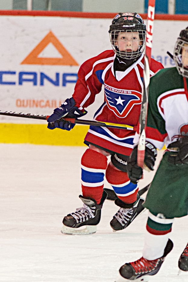 Zwei Personen in Sportkleidung und Helmen sind auf einem Eisfeld Schl├Ąger haltend unterwegs, mit einer Wand mit Werbung im Hintergrund.
