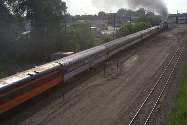 Ein Zug auf Schienen mit Bäumen, Strommasten mit Kabeln, Autos und Gebäuden zur Linken und einer Brücke vorne, aus der Rauch aufsteigt.