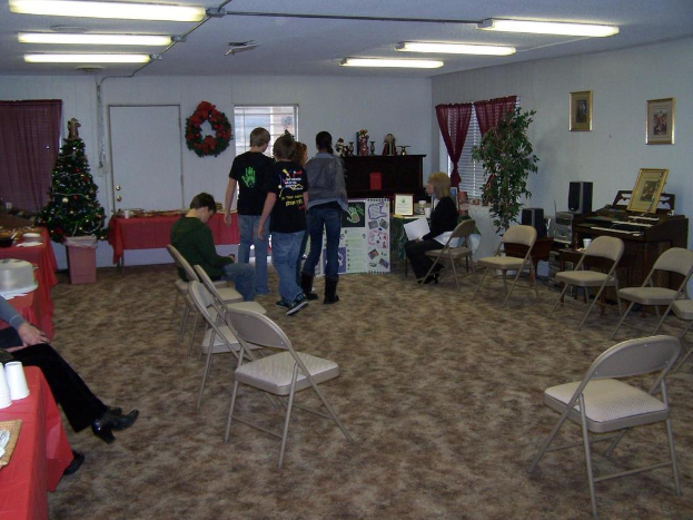 Ein Raum mit mehreren Stühlen, Tischen und Personen, einige stehen, einige sitzen, mit Tassen und Gefäßen auf den Tischen, einem Weihnachtsbaum, einem Piano auf der rechten Seite, Foto-Rahmen, Fenstern und Röhrenleuchten an der Decke und einer Wand im Hintergrund mit einem Strauß, Vorhang, Tür und weiteren Foto-Rahmen.