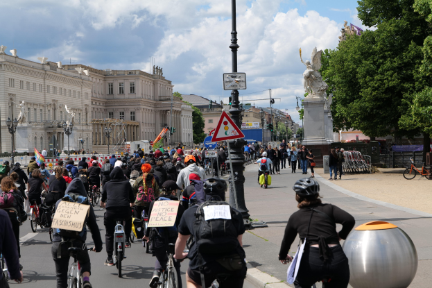Eine große Gruppe von Radfahrern fährt eine von Hochhäusern gesäumte Stadtstraße entlang, einige tragen Helme und haben Taschen dabei, andere halten Schilder; Pfosten mit Schildern und eine Statue sind im Hintergrund zu sehen, ebenso wie Bäume und ein bewölkter Himmel.