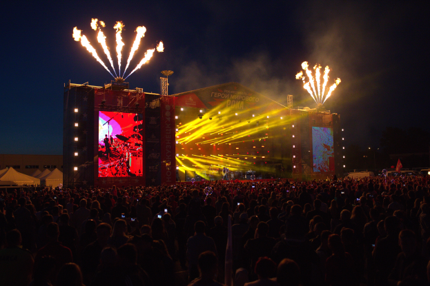 Eine große Menschenmenge versammelte sich auf einem Musikfestival, mit einer Bühne in der Mitte, auf der Musiker im Scheinwerferlicht spielten und von Fahnen umgeben waren, Zelte links und ein dunkler Himmel mit Feuerwerk im Hintergrund.