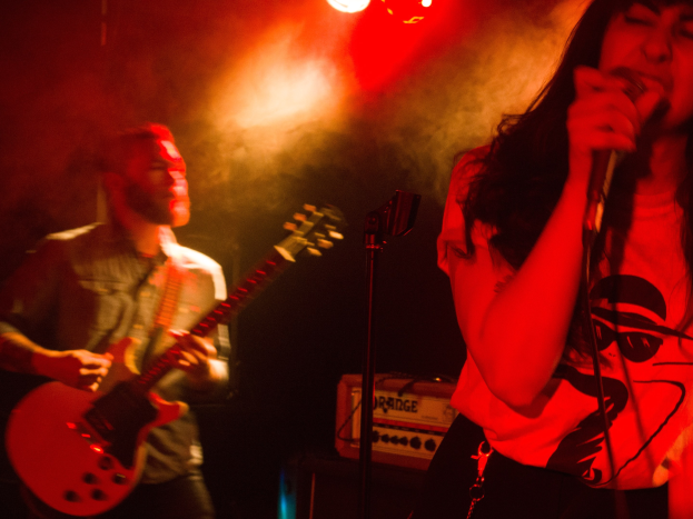Eine Frau, die in ein Mikrofon singt, auf einer Bühne mit einem Mann, der im Hintergrund eine Gitarre spielt, beleuchtet von Deckenlampen.