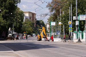 Eine Stadtstraße mit einer Baustelle, Fahrzeugen, Fußgängern, einem Radfahrer, Verkehrskegeln, Pfosten, Schildern, Strommasten mit Drähten, Bäumen, Gebäuden mit Fenstern und einer bewölkten Himmel.