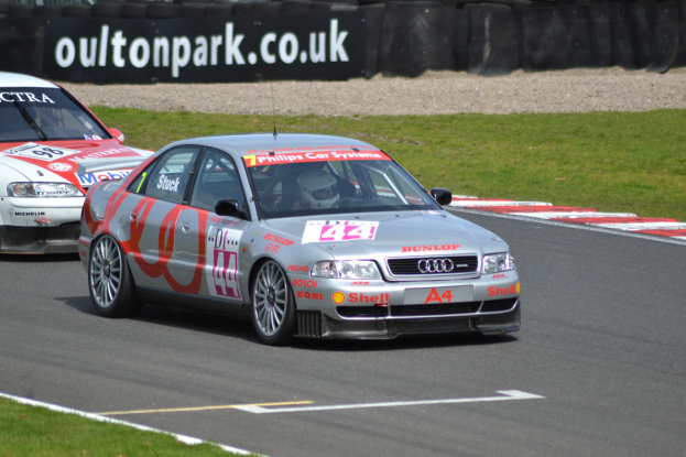 Zwei Autos auf einer grasumsäumten Rennstrecke mit Helmträgern im Hintergrund, umgeben von Reifen und einer Tafel mit Text.