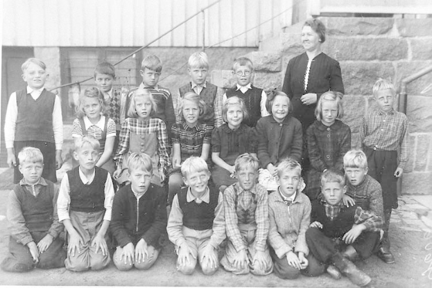 Schwarz-weiß-Foto einer Gruppe von Kindern, die vor einem Gebäude mit einer Wand und Treppen mit Geländern posieren, beschriftet mit "1930er Schulkinder" unten.