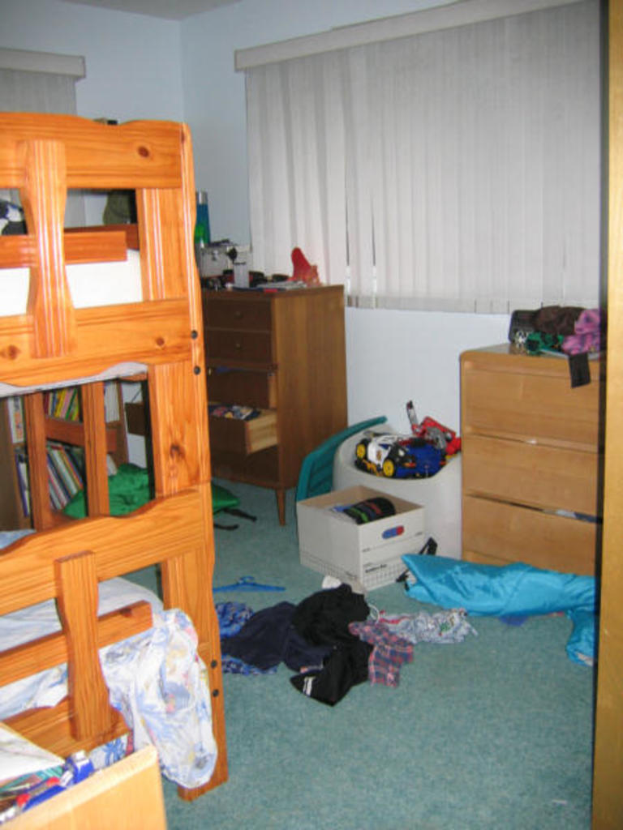 Ein Zimmer mit einem Stockbett, Schreibtisch, Tisch und verstreuten Kleidungsstücken und Gegenständen auf dem Boden.