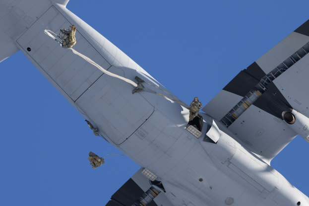 Ein großes weißes Militärflugzeug mit uniformierten Personen auf der Seite, das durch einen hellblauen Himmel fliegt.