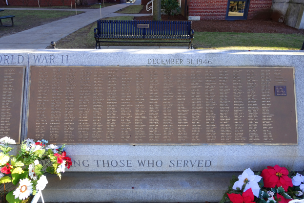 Ein Gedenkstein mit einer Bank davor, umgeben von Blumensträußen, mit Gebäuden, Bäumen, Pfählen und Fahrzeugen im Hintergrund.
