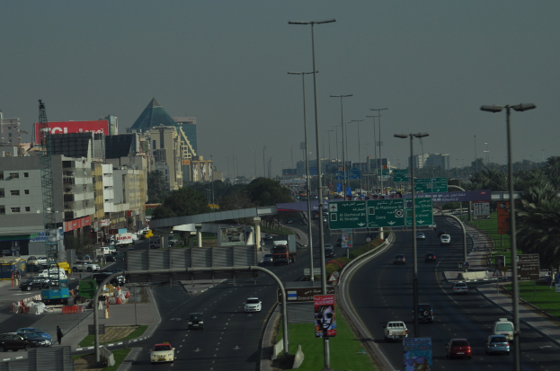 Eine Luftaufnahme einer Stadt mit Gebäuden, Türmen, Kraftfahrzeugen, Werbetafeln, Schildern, Straßen, Bäumen, Straßenlaternen, Menschen, Absperrgittern und Informationsanzeigen unter einem klaren Himmel.