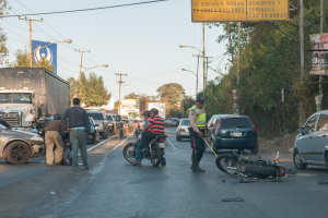 Eine Gruppe von Menschen steht um ein verunglücktes Motorrad auf der Straße herum, umgeben von mehreren Fahrzeugen, darunter ein Lkw, und einer Hintergrundlandschaft mit Bäumen, Pfählen, Lampen, Schildern und Himmel.