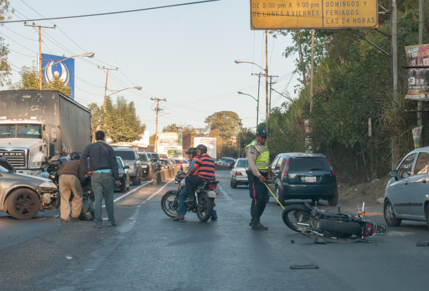 Eine Gruppe von Menschen steht um ein verunglücktes Motorrad auf der Straße herum, umgeben von mehreren Fahrzeugen, darunter ein Lkw, und einer Hintergrundlandschaft mit Bäumen, Pfählen, Lampen, Schildern und Himmel.