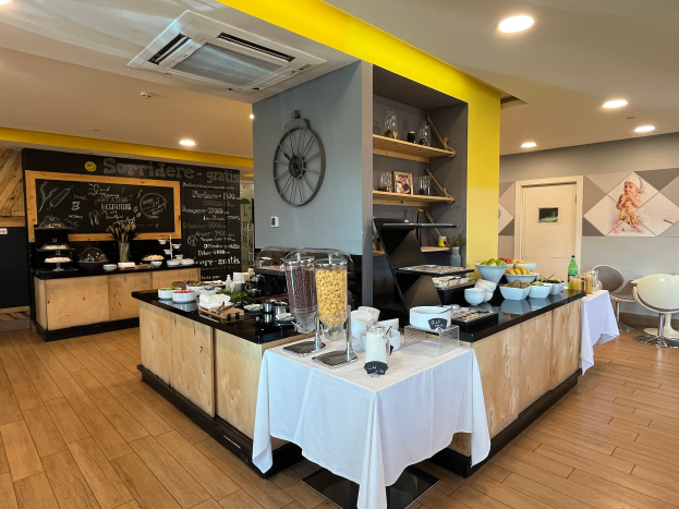 Ein Restaurantinnenraum mit Tischen und Stühlen, Lebensmitteln, einer Tafel mit Text, einer Wanduhr, Deckenlampen, einer Klimaanlage und einer Tür im Hintergrund, gelegen im Hôtel de Ville in Paris.