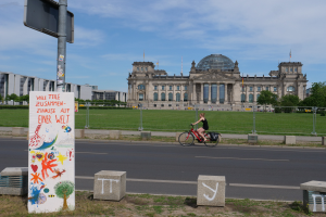 Eine Person in einem schwarzen Kleid fährt mit dem Fahrrad eine Straße vor dem Reichstag in Berlin, Deutschland, entlang, mit Gebäuden, Bäumen und ein paar Menschen im Hintergrund.