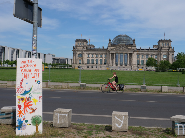 Eine Person in einem schwarzen Kleid fährt mit dem Fahrrad eine Straße vor dem Reichstag in Berlin, Deutschland, entlang, mit Gebäuden, Bäumen und ein paar Menschen im Hintergrund.