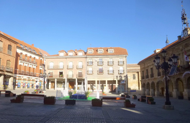 Ein Plaza Mayor auf einem Stadtplatz mit einem zentralen Brunnen, umgeben von Bänken, Topfpflanzen, Straßenlaternen, einem Uhrenturm und Gebäuden mit Fenstern unter einem klaren blauen Himmel.