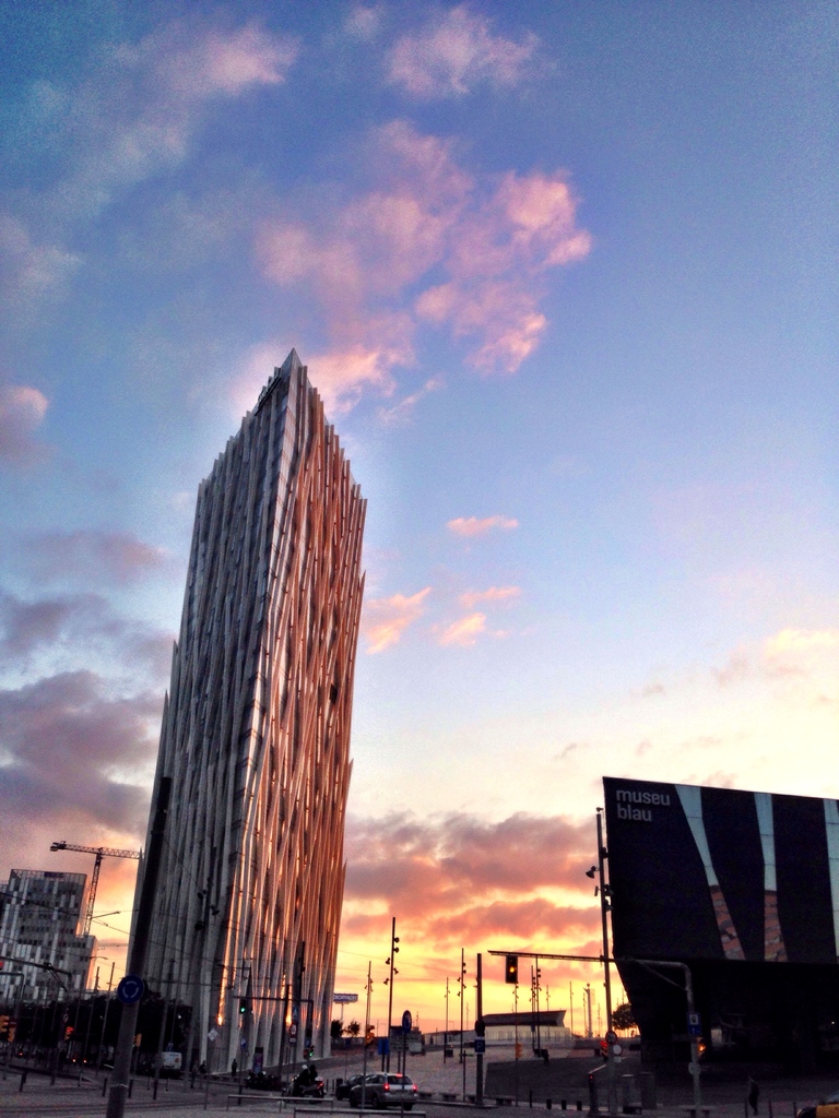 Eine Stadtansicht mit einem prominenten Wolkenkratzer, zahlreichen Gebäuden, einer Reklamewand rechts, einer Straße mit einem Auto unten und Wolken am Himmel darüber.