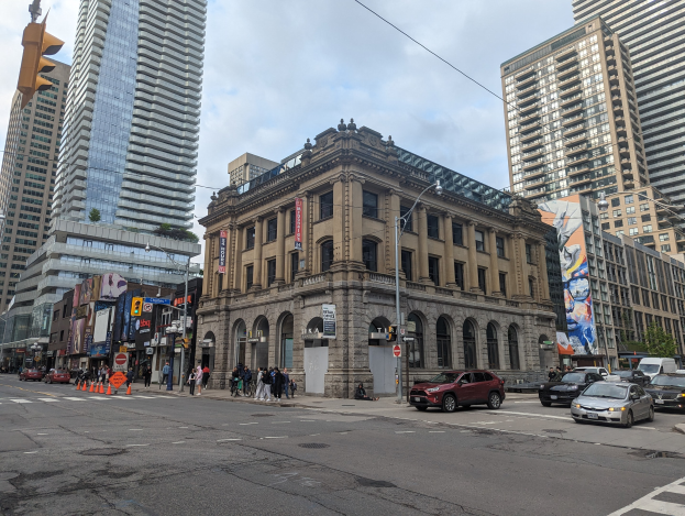 Eine belebte Stadtstraße mit hohen Gebäuden, Fahrzeugen, Fußgängern, Verkehrskegeln, Laternen, Verkehrsampeln, Plakaten, Werbetafeln, Bäumen und der Toronto Public Library als Mittelpunkt unter einem bewölkten Himmel.