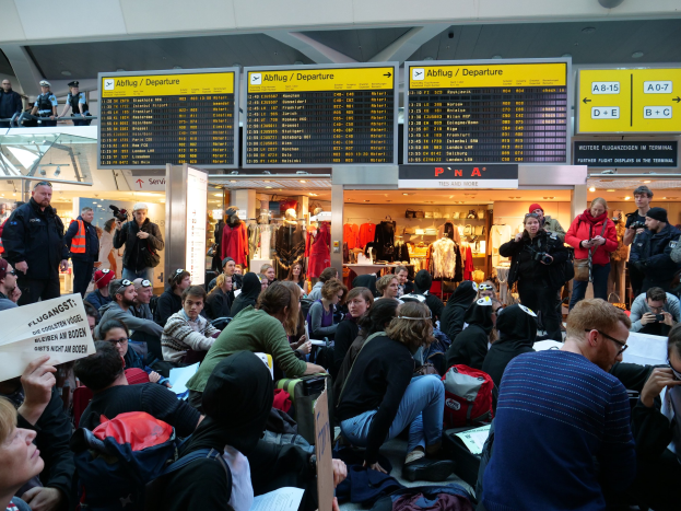 Eine große Gruppe von Menschen in einem Flughafen, einige sitzen mit Taschen und Papieren, andere stehen, mit Schildern, Schaufensterpuppen in Kleidern und Deckenbeleuchtung im Hintergrund, was auf eine Demonstration hinweist.