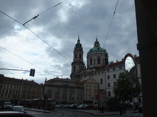 Gebäude in der Mitte, ein Pfahl und Bäume auf der rechten Seite, Autos, Menschen und ein Zug unten und Drähte oben.