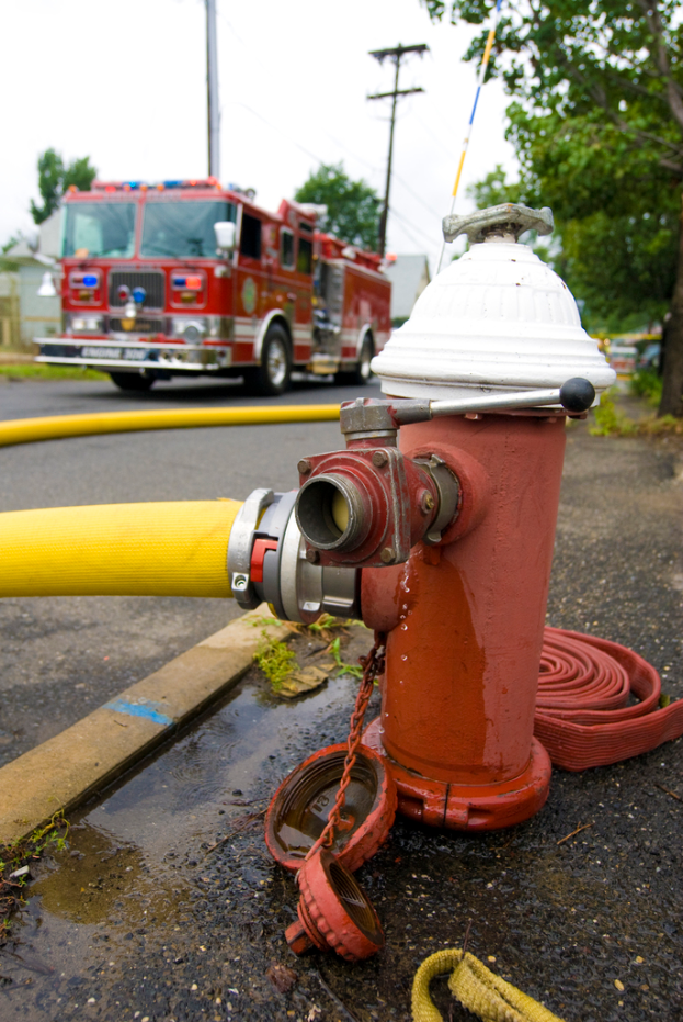 Ein Feuerhydrant und ein gelber Schlauch vorne mit einem Bus, Stromleitungen, Masten und Bäumen im Hintergrund.