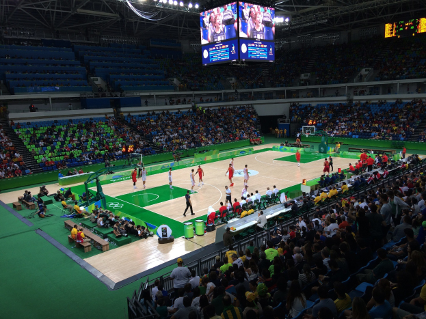 Große Halle mit Zuschauern auf Sitzen und Stehplätzen rund um einen Basketballplatz, Überwachungsbildschirme und Beleuchtung sowie ein digitales Anzeigebrett auf der rechten Seite.