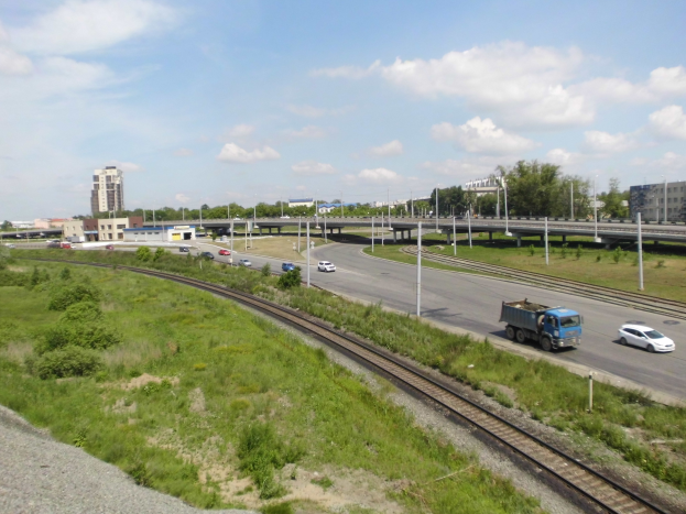 Eine Bahnschiene verläuft durch eine Stadtlandschaft mit Fahrzeugen, Grünflächen, Polen, Brücken, Gebäuden und einem bewölkten Himmel.